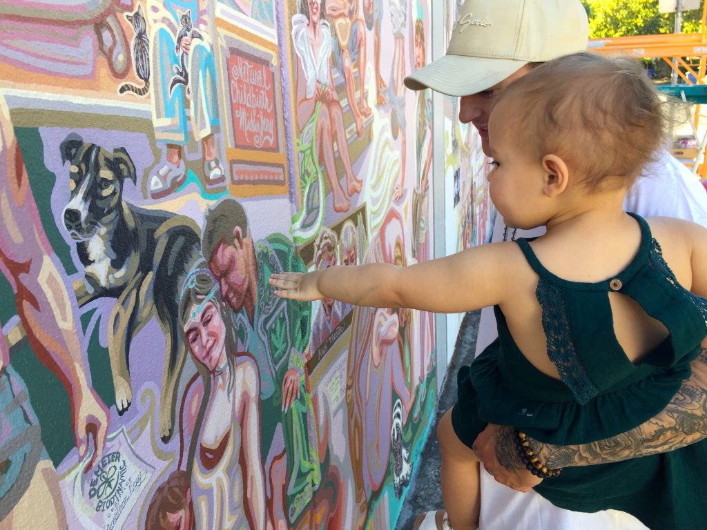 A father holds his toddler who reaches out to touch the mural